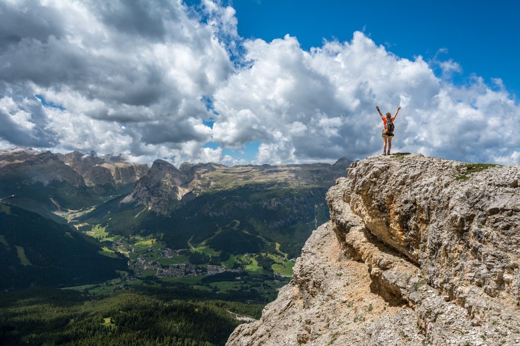 jedna osoba stojąca na szczycie w uniesionymi rozłożonymi rękami, w tle góry